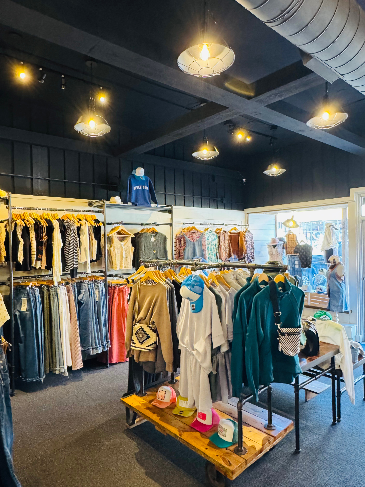 The boutique floor with denim, tops, and a display cart of trucker hats under black pendant lighting